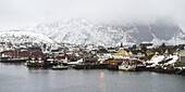 Gebäude und Boote am Wasser unter bewölktem Himmel mit schneebedeckten Bergen; Lofoten-Inseln, Nordland, Norwegen