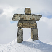 A Stone Inuksuk In The Snow Under A Cloudy Sky With A Snow Covered Mountain In The Background; Whistler, British Columbia, Canada