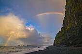 Die als Reynisdrangar bekannten Meeresfelsen mit Wellen bei Sonnenaufgang und Tausenden von Vögeln, die mit einem Regenbogen durch die Luft fliegen, Südküste; Island