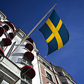 Niedriger Blickwinkel auf die schwedische Flagge und ein Gebäude mit roten Markisen über den Fenstern, Ostermalm District; Stockholm, Schweden