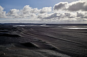 Ein Fluss fließt durch ein Flussbett aus schwarzem Sand mit Bergen in der Ferne; Island
