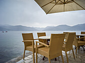 Tische und Stühle unter einem Sonnenschirm an der Uferpromenade der Bucht von Kotor; Bjelila, Opstina Tivat, Montenegro.