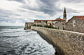 A stone wall lining the shoreline with buildings and a church tower; Budva, Opstina Budva, Montenegro