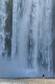 Frau steht am Fuße des Skogafoss-Wasserfalls und schaut auf das kraftvoll fließende Wasser; Island