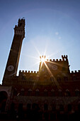 Von hinten beleuchteter Palazzo Pubblico (Rathaus) und Mangia-Turm mit Sonnenaufgang und blauem Himmel; Siena, Toskana, Italien.
