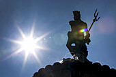 Silhouette of statue on top of fountain with sunburst and blue sky; Trento, Trento, Italy