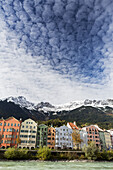 Bunte Gebäude entlang des Flussufers mit schneebedeckten Berggipfeln, dramatischen Wolken und blauem Himmel über der Stadt; Innsbruck, Tirol, Österreich.