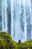 Mann sitzt auf einem moosbewachsenen Felsen am Fuße des Skogafoss-Wasserfalls; Island