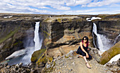 Eine junge asiatische Wanderin posiert für ein Porträt am Rande einer atemberaubenden Landschaft mit zwei Wasserfällen, die als Haifoss bekannt ist; Island