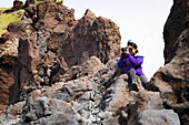 A tourist sits on the top of a rock formation to take a higher photo of the landscape; Iceland