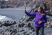 Eine Touristin posiert für ein Selbstporträt mit ihrem Handy am schwarzen Sandstrand in Westisland, Snaefellsnes-Halbinsel; Island.