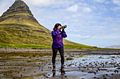 Eine Wanderin hält bei Ebbe am Strand an, um ein Foto mit einer Spiegelreflexkamera vor dem Berg Kirkjufell auf der Halbinsel Snaefellsnes im Westen Islands zu machen; Island