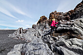 A female hiker climbs a lava rock formation on the black sand beach in Western Iceland; Iceland