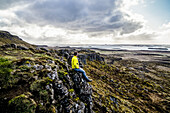 Mann blickt über den Ozean, während er auf einer Klippe sitzt; Island