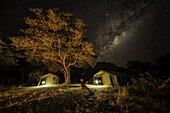 Die Milchstraße am Himmel, darunter ein Zelt in einem Buschcamp, während ein Mann sitzt und in den Himmel schaut; Botswana
