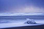 Piece of ice being hit by waves along the South shore of Iceland with stormy skies behind it; Iceland