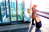 A woman stands with her suitcase on the platform of a train station beside the tracks and uses her smart phone; St. Gallen, St. Gallen, Switzerland