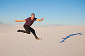 Carefree man in mid-air on the white sand with blue sky, casting a shadow beside him, White Sands National Monument; Alamogordo, New Mexico, United States of America