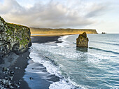 Cliffs and a rock formation along the coastline of the Southern Region of Iceland, with the surf washing up onto black sand in the foreground; Myrdalshreppur, Southern Region, Iceland