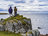 A man and woman tourist stand on a rocky ridge overlooking the ocean and coastline in Northern Iceland; Hunathing vestra, Northwestern Region, Iceland