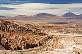 Straße führt hinunter in ein hochgelegenes Wüstental mit einzigartigen Felsformationen auf der linken Seite und einem Vulkangipfel in der Ferne; San Pedro de Atacama, Antofagasta, Chile