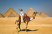 Weibliche Touristin winkt, während sie auf einem Kamel sitzt, Pyramidenkomplex von Gizeh, UNESCO-Welterbe; Gizeh, Ägypten.