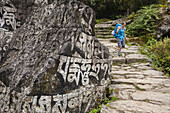 A woman wearing a backpack walks down a stone trail, pat a rock wall with script on it; Nepal