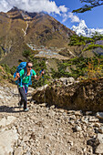 Woman trekker climbing up the rocky trail towards Mong La with the village of Phortse seen on a Himalayan mountain terrace across the valley and Ama Dablam rising up in the background on a sunny autumn day in Sagarmatha National Park;  Solokhumbu District, Nepal