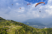 Eine Gruppe farbenfroher Gleitschirmflieger spielt in der Thermik über den Terrassen von Sarangkot mit der Stadt Pokhara in der Ferne, an einem sonnigen Herbsttag mit bewölktem Himmel; ?Kaski Distrikt, Nepal