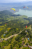 Luftaufnahme einer Gruppe von Gleitschirmfliegern, die über Sarangkot fliegen und in der Thermik spielen, mit der Stadt Pokhara und dem Phewa-See in der Ferne, an einem bewölkten Herbsttag; Pokhara, Nepal.