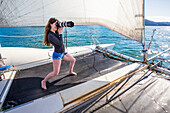 Woman standing on a catamaran taking photographs with a camera with a telephoto lens on a boat tour through the Abel Tasman National Park; Tasman, South Island, New Zealand