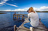 View taken from behind of a woman taking a photograph of another woman on a wooden dock while exploring Lake Mahinapua; West Coast, New Zealand