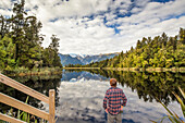 Tourist explores the Lake Matheson Mirror Lakes. The cloudy sky somewhat dilutes the reflection on this day; South Westland, New Zealand