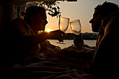 A young couple cheers with wine glasses in the back of a camper van at sunset. A group of travellers pull up alongside a Bratislava lake to free camp for the night; Bratislava, Bratislava Region, Slovakia