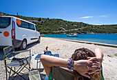 Frau sitzt in der Sonne und ruht sich außerhalb eines Wohnmobils aus, als eine Gruppe von Reisenden in Slano für einen Nachmittag am Strand anhält; Slano, Gespanschaft Dubrovnik-Neretva (Dubrova?ko-neretvanska ?upanija), Kroatien