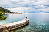 Touristen spazieren auf einem Steg in Richtung des türkisfarbenen Wassers an der Küste von Podgora, Kroatien. Diese Gruppe hat nach dem Campen in ihrem Wohnwagen eine Pause eingelegt, um frühmorgens zu schwimmen und zu duschen; Podgora, Gespanschaft Split-Dalmatien, Kroatien