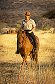 Frau reitet auf einem Pferd (Equus ferus caballus) durch den Busch in der Savanne im warmen Licht des Sonnenuntergangs, schaut in die Kamera und lächelt auf der Gabus Game Ranch; Otavi, Otjozondjupa, Namibia.