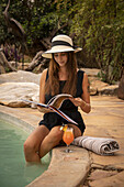 Frau mit Sonnenhut sitzt an einem Swimmingpool und liest auf der Gabus Game Ranch; Otavi, Otjozondjupa, Namibia.