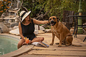 Frau mit Sonnenhut sitzt an einem Swimmingpool und streichelt einen Hund (Canis lupus familiaris) auf der Gabus Game Ranch; Otavi, Otjozondjupa, Namibia.