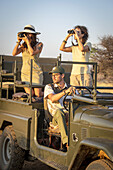 Nahaufnahme von Frauen in einem Jeep, eine mit einem Fernglas und eine mit einer Kamera, die in die Savanne blicken, während der Fahrer hinter dem Lenkrad sitzt, während einer Safari auf der Gabus Game Ranch; Otavi, Otjozondjupa, Namibia.
