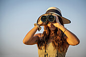 Nahaufnahme einer Frau, die einen Strohhut trägt und durch ein Fernglas in das helle Sonnenlicht vor dem blauen Himmel auf der Gabus Game Ranch blickt; Otavi, Otjozondjupa, Namibia.