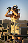 Nahaufnahme einer Frau, die in einem Jeep auf Safari kniet, einen Strohhut trägt, eine Kamera um den Hals hat und mit einem Fernglas auf die Savanne der Gabus Game Ranch bei Sonnenuntergang schaut; Otavi, Otjozondjupa, Namibia.