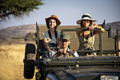 Women sitting in the back seat of a jeep on safari with a guide driving them through the savanna, looking at camera at the Gabus Game Ranch; Otavi, Otjozondjupa, Namibia