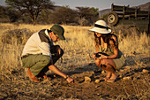 Safari guide with woman traveler wearing a straw hat and holding a camera crouching down on the ground talking on the savanna at the Gabus Game Ranch at sunset; Otavi, Otjozondjupa, Namibia