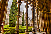 Looking out through the ornate columns of the cloister into the formal garden of the inner courtyard towards the chapel at the medieval Monastery of Batalha, a masterpiece of Gothic architecture; Batalha, District of Leiria, Centro Region, Portugal