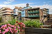 From balconies with flower boxes looking across the Tsavkisi-Tskali River to the old brick buildings with wooden balconies built on the cliffs in Legvtakhevi with the minaret of the Tbilisi Mosque in the background, part of the historical neighborhood of Abanotubani in the Old Town; Tbilisi, Georgia
