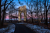 George Washington Bridge (specially lit for Martin Luther King Jr. Day) seen through the silhouetted bare trees along the pathway at the Fort Lee Historic Park at dusk; Fort Lee, New Jersey, United States of America