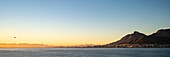 Silhouette eines Hubschraubers, der über die Tafelbucht in Richtung des Küstenhafens und des Hafens von Kapstadt fliegt, mit Blick auf Devil's Peak und den Tafelberg in der Morgendämmerung vom Meer aus; Kapstadt, Kapprovinz, Südafrika.