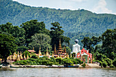 Blick von hinten auf einen Riesenbuddha neben Pagoden und goldenen Stupas auf der Insel Shwe Paw und Booten, die entlang des Ayeyarwady (Irrawaddy) Flusses vertäut sind; Shwegu, Kachin, Myanmar (Burma)