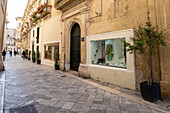 Straßenszene mit Menschen, die an Schaufenstern und Kalksteingebäuden in der Altstadt von Lecce vorbeigehen; Lecce, Apulien, Italien.